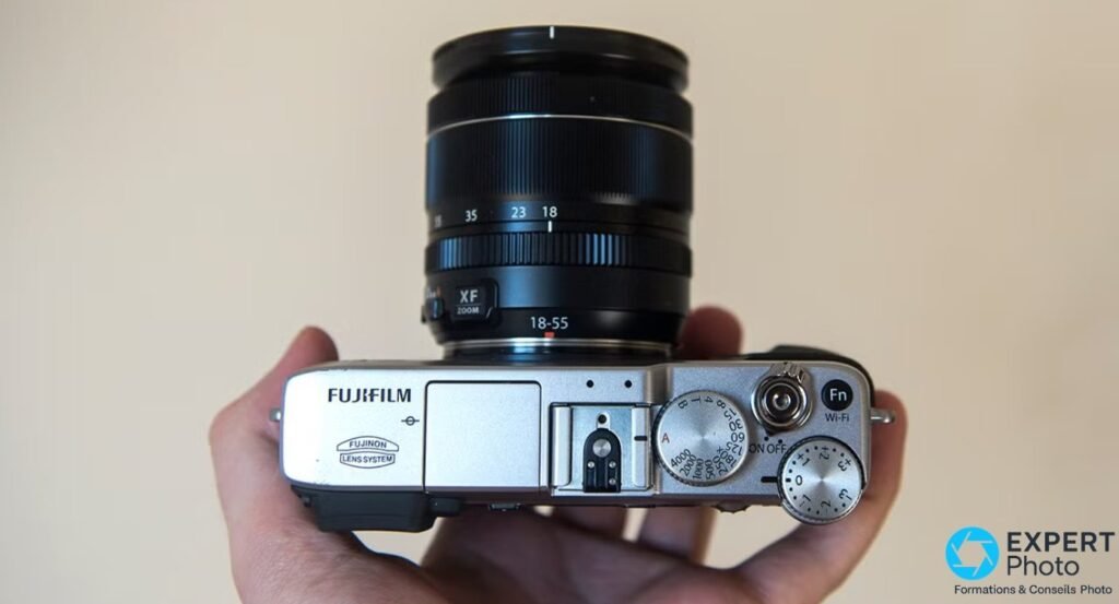 Vue de dessus du Fujifilm X-E2 tenu en main, molettes vitesses et correction d'expo, bouton Fn Wi-Fi, optique XF 18-55mm. 