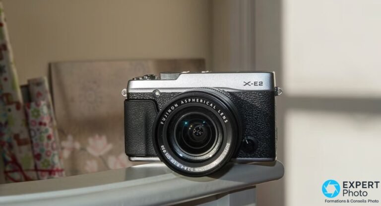 Fujifilm X-E2 silver avec Fujinon XF 18-55mm f/2.8-4 R LM OIS, posé sur une étagère blanche en lumière de fenêtre.