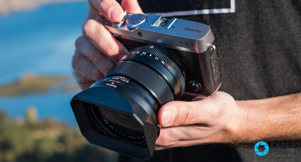 Fujifilm X-E3 tenu à deux mains avec un Fujinon XF 16mm f/1.4 R WR et pare-soleil tulipe, en extérieur face à un plan d'eau. 