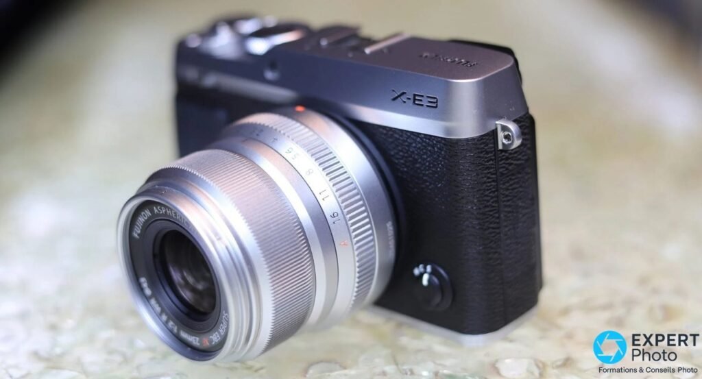 Fujifilm X-E3 silver et noir avec Fujinon XF 23mm f/2 R WR silver, vue 3/4 avant gauche, posé sur surface granit beige floue. 