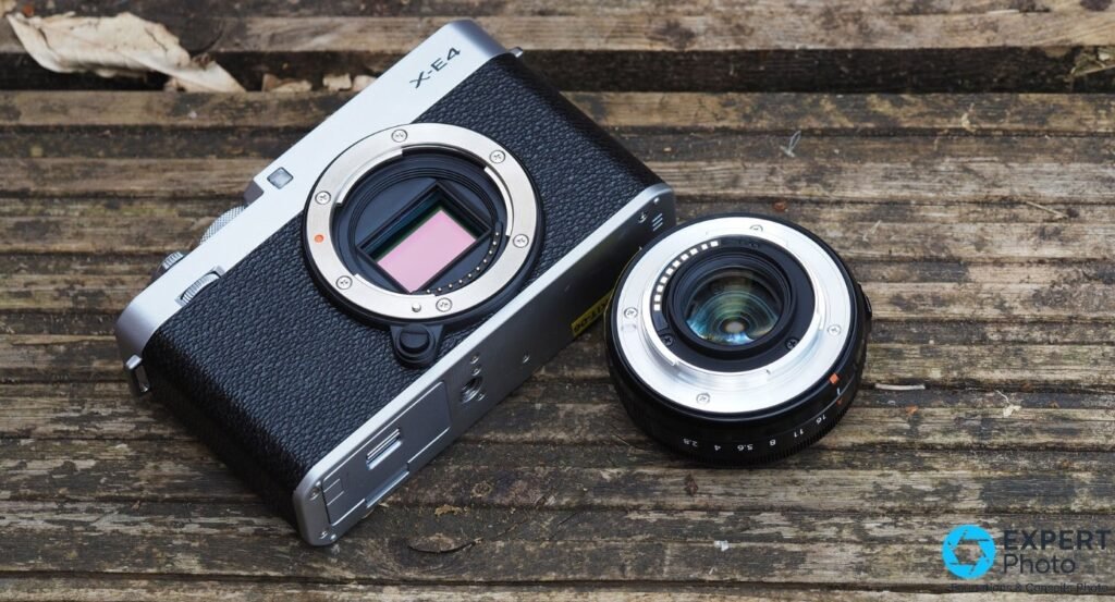 Fujifilm X-E4 argent et noir avec XF 27mm f/2.8 sans cache, vue 3/4 avant gauche posé sur mur en pierre, fougères en fond.