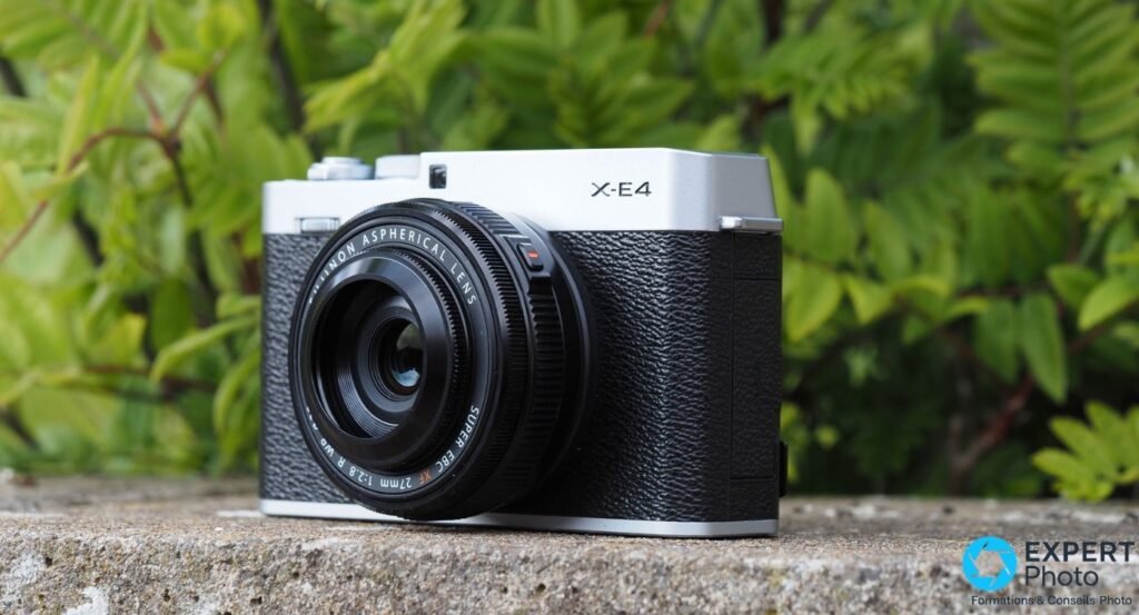 Fujifilm X-E4 argent et noir avec XF 27mm f/2.8, vue 3/4 avant sur rebord en terre cuite, fleurs roses en arrière-plan.