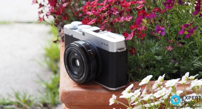 Test Fujifilm X-E4 : un APS-C compact qui tient encore en 2026 Fujifilm X-E4 argent et noir avec XF 27mm f/2.8 monté, vue 3/4 avant centrée, feuillage vert flou en arrière-plan.