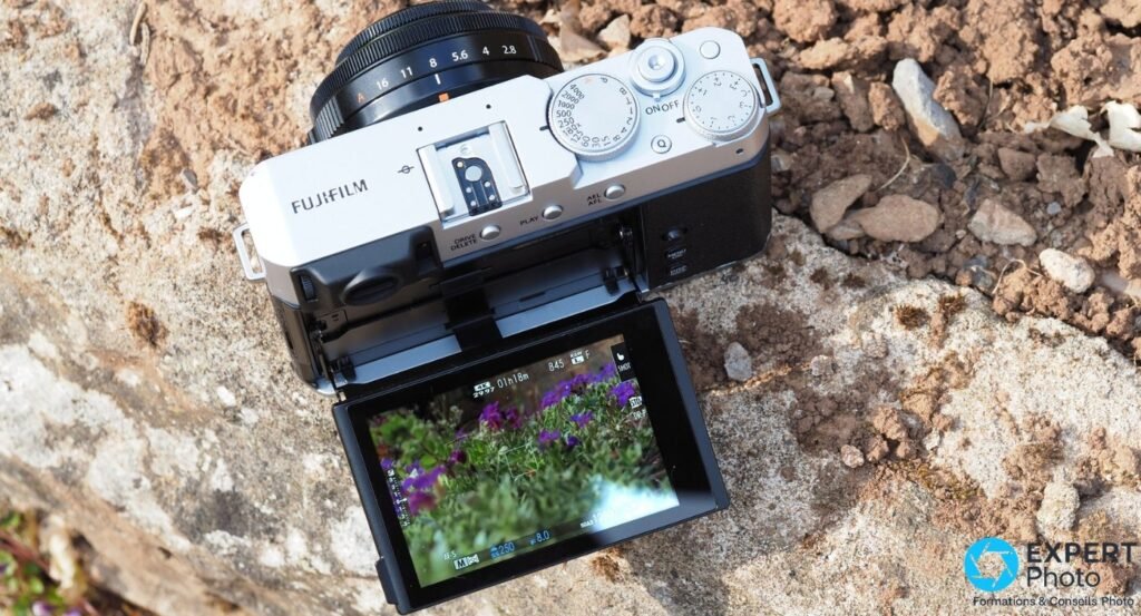 Fujifilm X-E4 vu de dessus avec écran tactile basculé 180° vers l'avant en live view, posé sur mobilier de jardin.