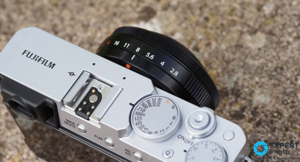 Fujifilm X-E4 boîtier nu, monture X et capteur APS-C visibles, XF 27mm f/2.8 démonté posé à côté sur planches de bois.