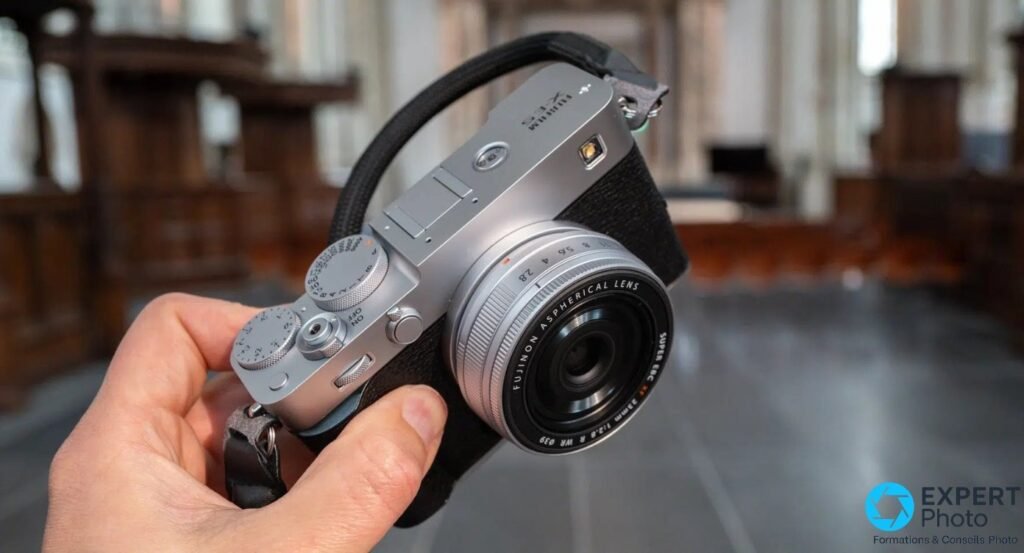 Fujifilm X-E5 silver tenu en main, vue trois-quarts dessus, molettes et objectif XF 23mm F2.8 visibles.