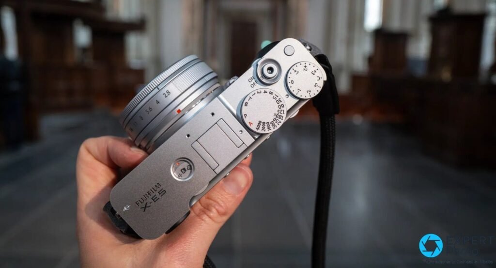 Fujifilm X-E5 silver tenu en main, vue de dessus, molettes de vitesse et compensation d'exposition visibles.