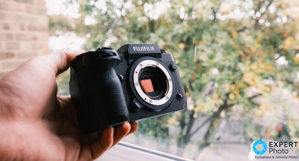 Boîtier Fujifilm X-H2 nu vu de face, capteur APS-C orange visible dans la monture X, tenu d'une main.