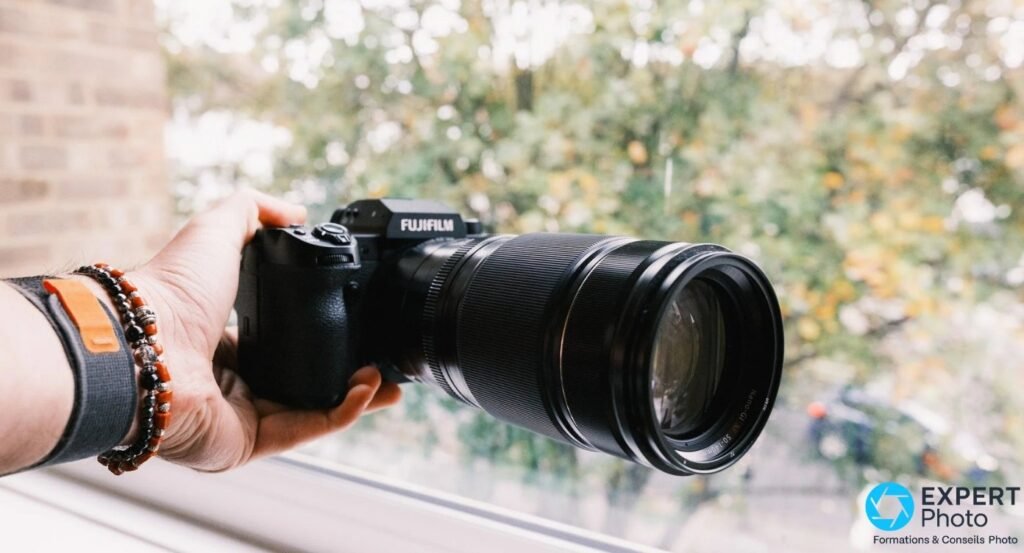 
Boîtier Fujifilm X-H2 noir avec téléobjectif zoom monté, tenu en main devant une fenêtre, logo FUJIFILM lisible. 
