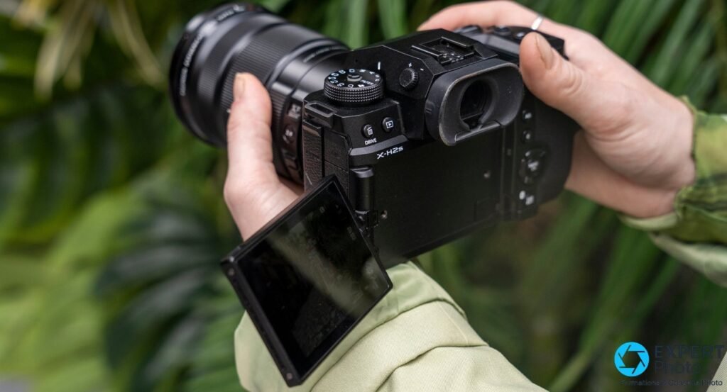 Fujifilm X-H2S tenu à deux mains, objectif noir monté, écran arrière articulé déployé vers le bas