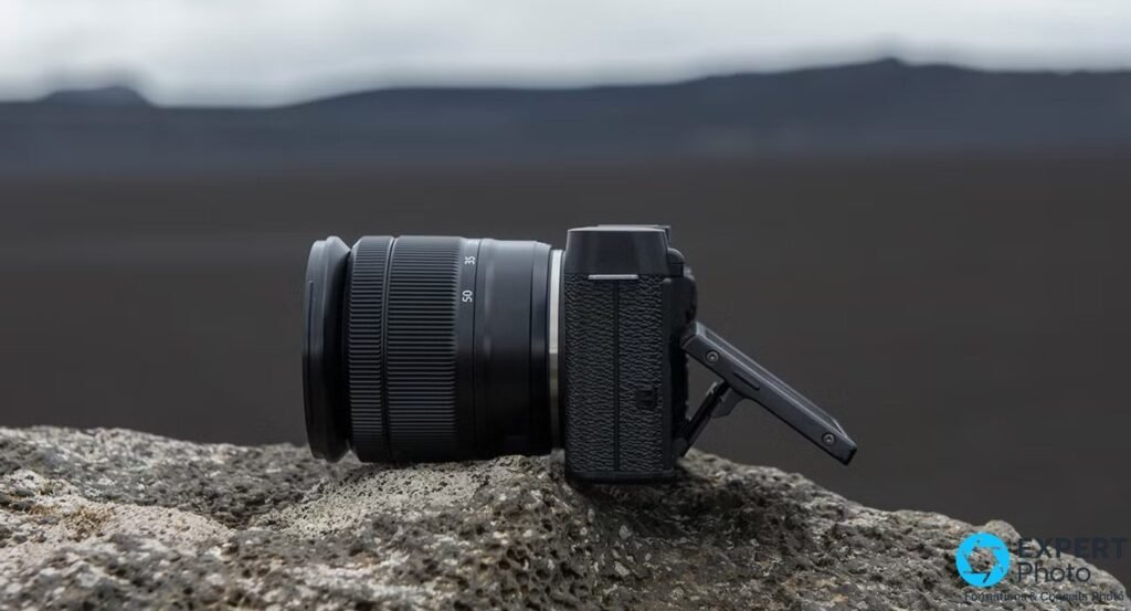 Fujifilm X-M1 noir vu de profil gauche, écran arrière inclinable déployé sur charnière, posé sur une roche, paysage flou