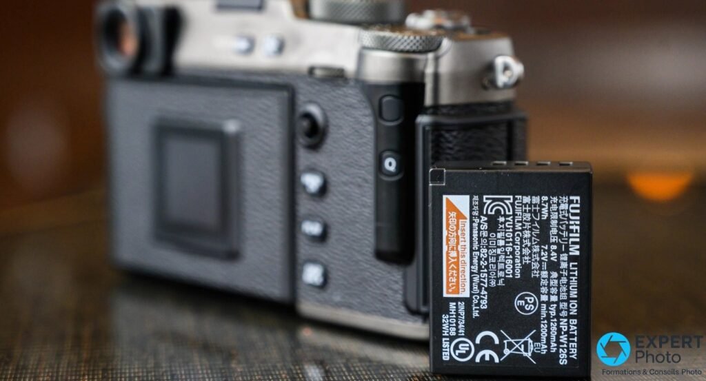 Batterie Fujifilm NP-W126S nette au premier plan, dos d'un Fujifilm X-Pro3 finition argent flou en arrière-plan.