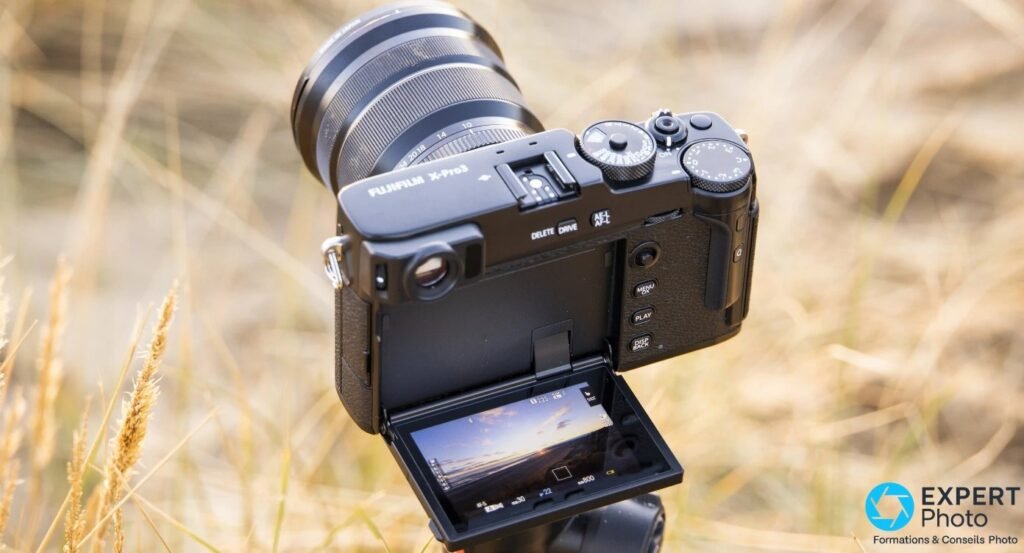 Fujifilm X-Pro3 noir en plongée, écran principal déplié vers le bas affichant un live view de paysage, fond herbes sèches.