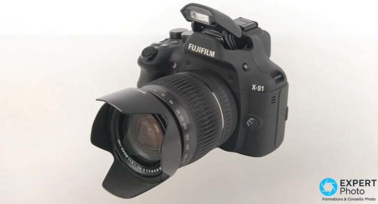 Fujifilm X-S1 vu de 3/4 avant, flash pop-up sorti, zoom 24-624 mm avec pare-soleil et échelle focales visibles.