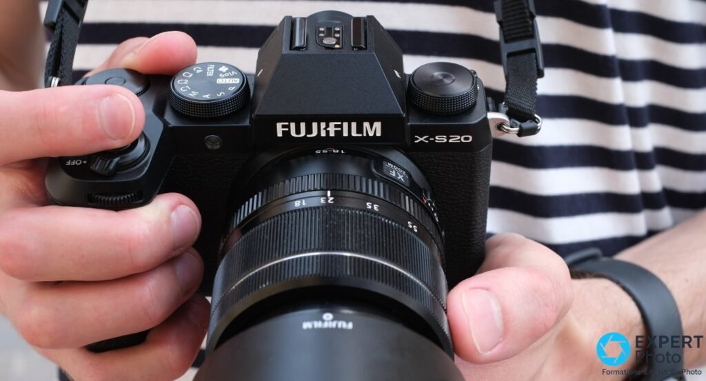 Fujifilm X-S20 tenu à deux mains avec objectif XF 18-55mm et pare-soleil monté, en position de prise de vue