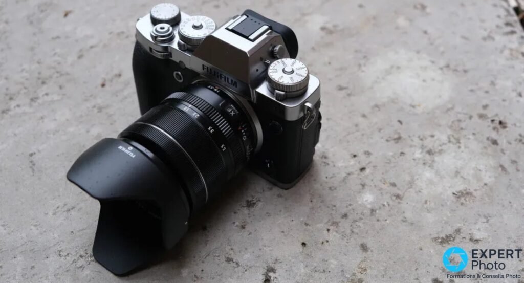 Fujifilm X-T3 silver équipé d'un objectif XF 18-55mm avec pare-soleil pétale, vu de trois quarts avant sur surface béton