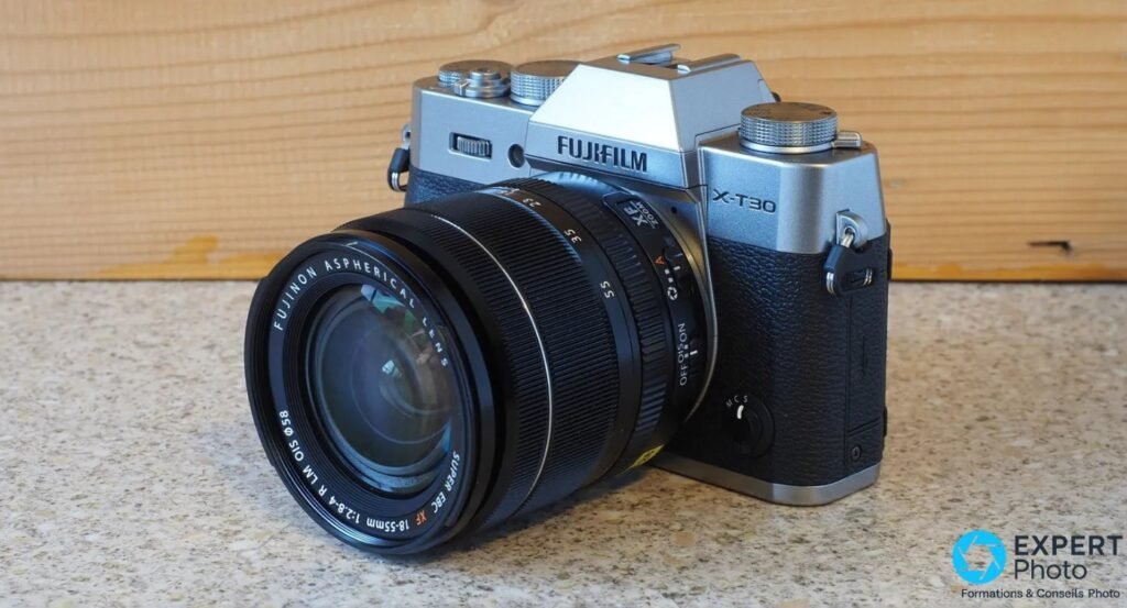 Fujifilm X-T30 II silver équipé du Fujinon XF 18-55mm f/2.8-4 OIS, vue 3/4 avant gauche, surface beige et fond bois.