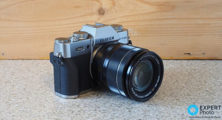 Test Fujifilm X-T30 II : un APS-C à molettes encore défendable Fujifilm X-T30 II silver équipé du Fujinon XF 18-55mm f/2.8-4, vue 3/4 avant sur surface beige et fond bois.