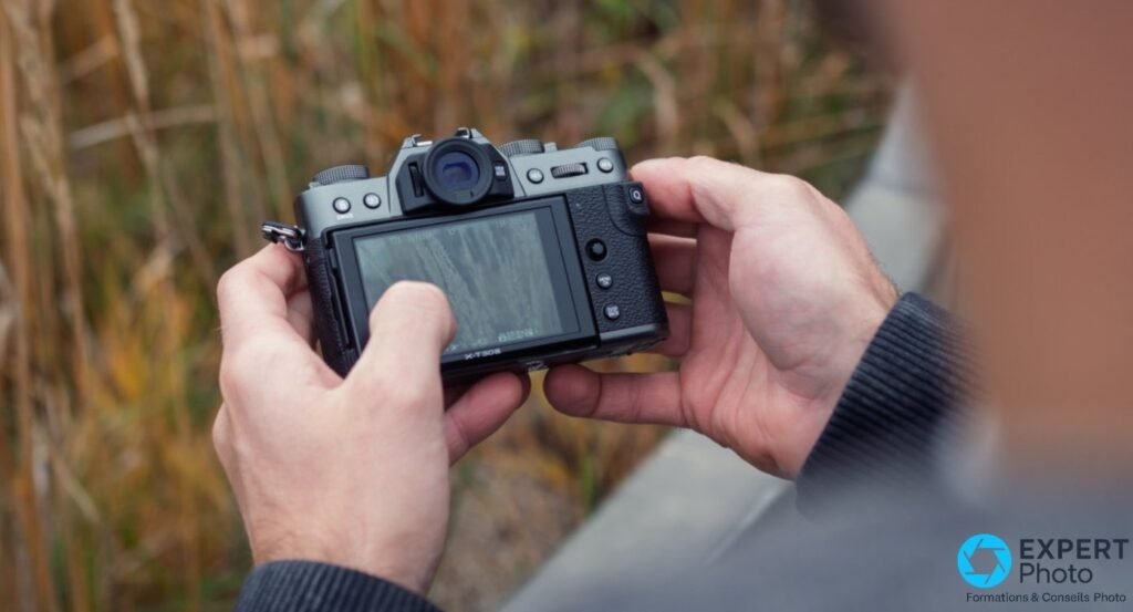 Fujifilm X-T30 III noir tenu à deux mains, vue arrière avec écran allumé en live view sur fond d'herbes hautes.