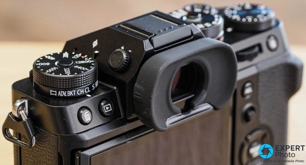 Gros plan sur les molettes ISO et drive du Fujifilm X-T5, œilleton EVF, hot shoe et boutons trash visibles.