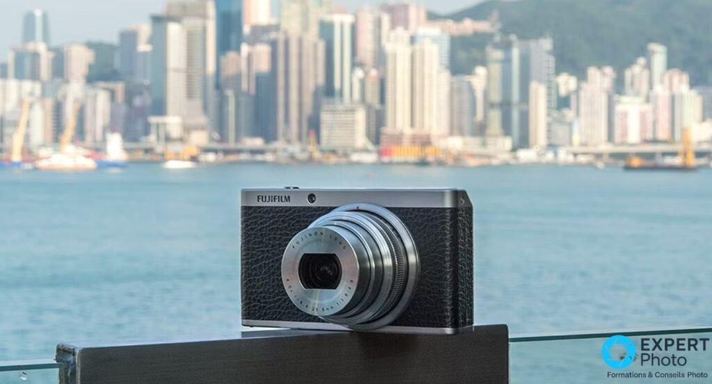 Fujifilm XF1 noir et argenté, zoom déployé, posé sur rambarde devant skyline urbaine floue