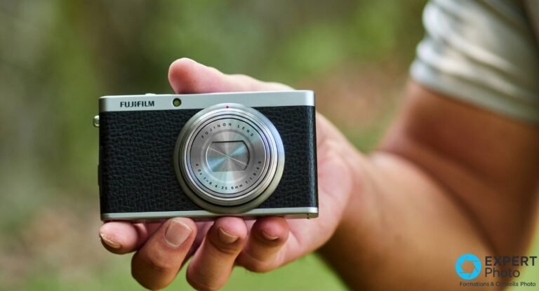 Fujifilm XF1 noir et argenté tenu dans une main devant fond végétal flou, optique rétractée