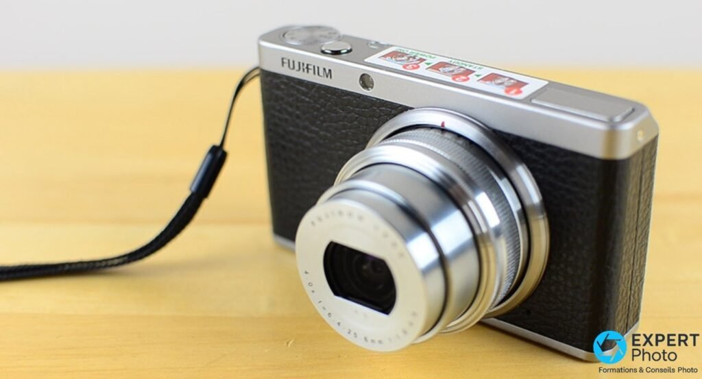 Fujifilm XF1 vu de 3/4 avant avec zoom mécanique déployé et dragonne tressée noire, posé sur bois clair