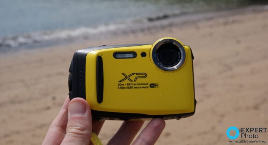 Fujifilm XP130 jaune tenu dans une main, vue de face, dragonne visible, plage de sable et mer floue en arrière-plan