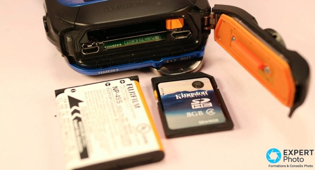Compartiment batterie du Fujifilm XP70 ouvert, joint étanchéité orange visible, batterie NP-45S et carte SD Kingston posées 