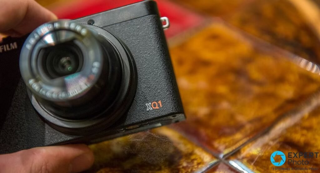 Fujifilm XQ1 vu de trois quarts avant, objectif déployé légèrement flou, logo XQ1 orange visible sur le boîtier noir.