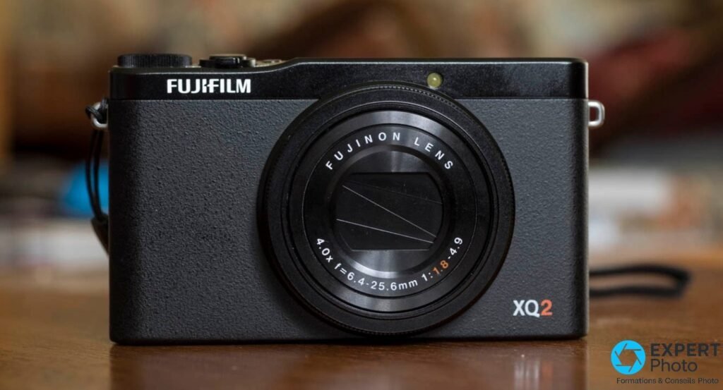 Fujifilm XQ2 noir vu de face, objectif Fujinon LENS rétracté, posé sur surface bois, sangle visible.