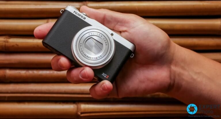 Test Fujifilm XQ2 : un compact expert encore pertinent en occasion ? Fujifilm XQ2 silver tenu en main droite, objectif Fujinon LENS déployé en grand-angle, fond bambou flou.