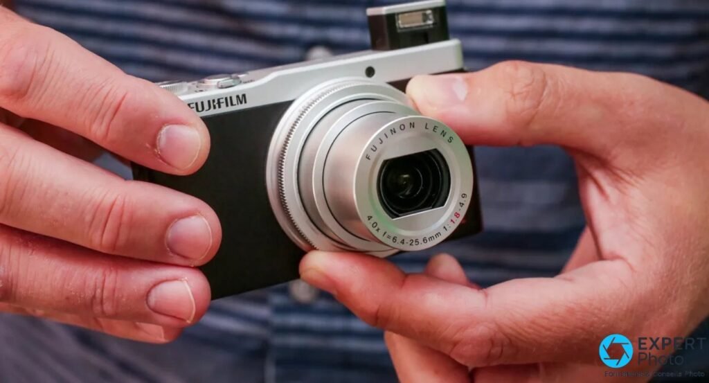 Fujifilm XQ2 silver tenu à deux mains, flash pop-up sorti, objectif Fujinon LENS déployé en téléobjectif.