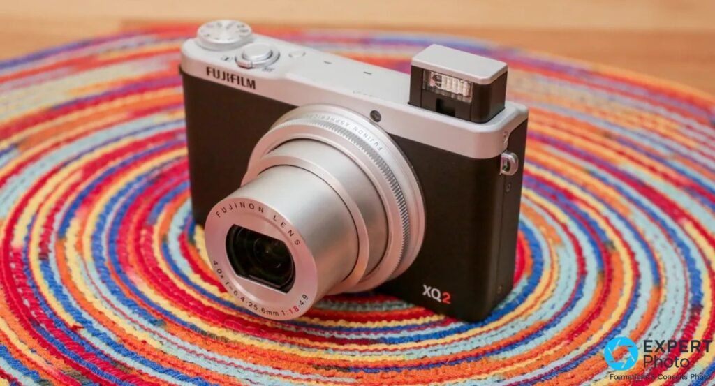 Fujifilm XQ2 silver vu de trois-quarts, flash pop-up sorti, objectif déployé, posé sur tapis coloré.