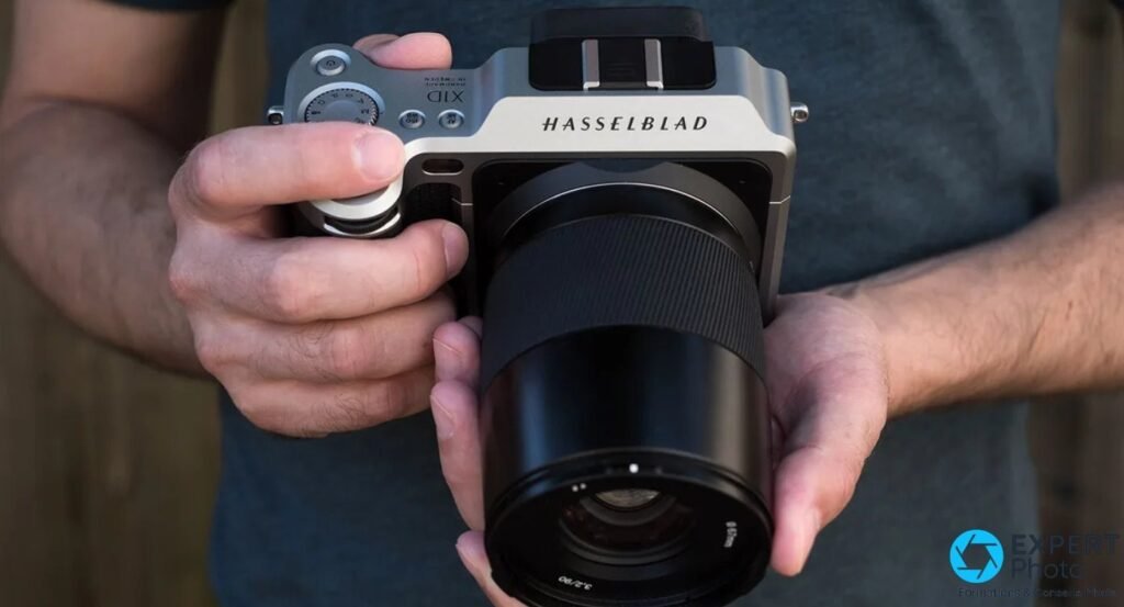Hasselblad X1D-50c argent tenu à deux mains, objectif XCD noir, sur fond de t-shirt gris flou