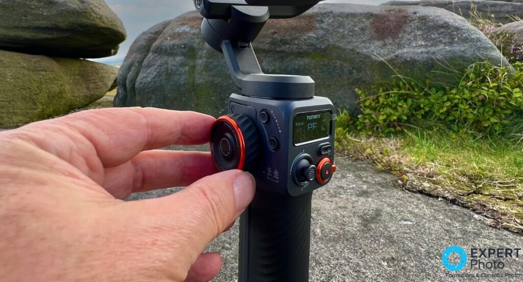 Gimbal Hohem iSteady M6 Kit tenu en main, pouce sur la molette à anneau rouge, écran OLED affichant FOCUS PF