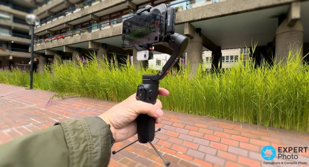 Gimbal Hohem iSteady V3 déplié tenu à bout de bras, mini-trépied ouvert, smartphone monté, fond urbain flou