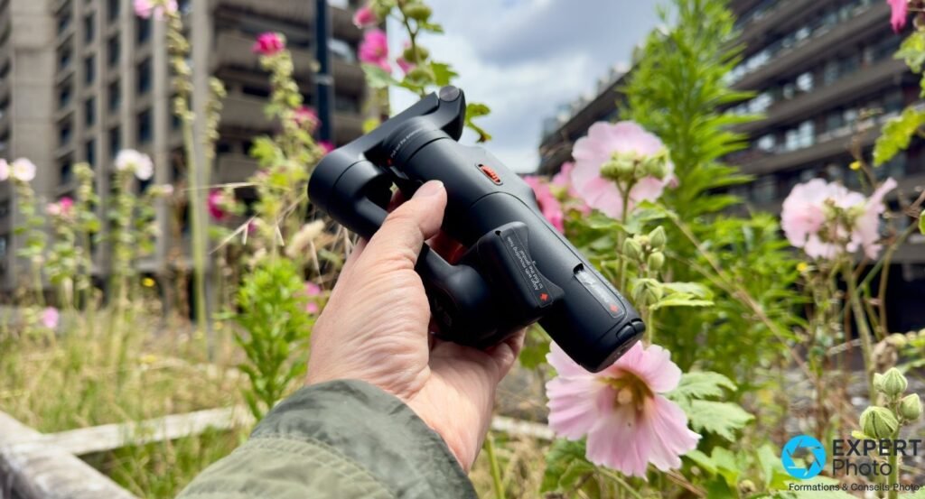 Hohem iSteady V3 plié tenu en main, tête repliée vers le bas, fleurs roses et bâtiments flous en arrière-plan
