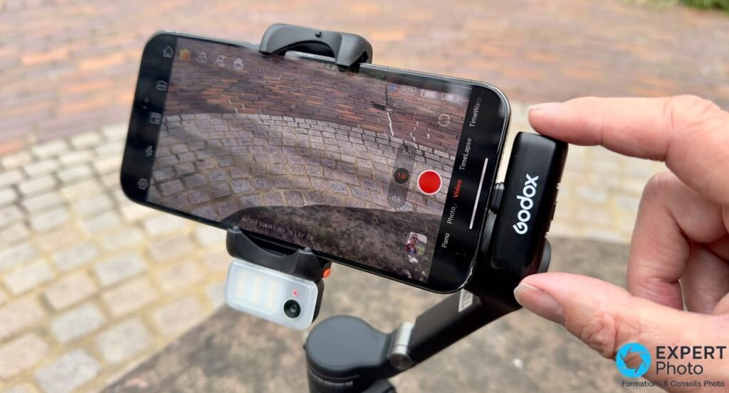 Hohem iSteady V3 avec smartphone filmant des pavés, émetteur Godox clipsé et module LED sous le téléphone