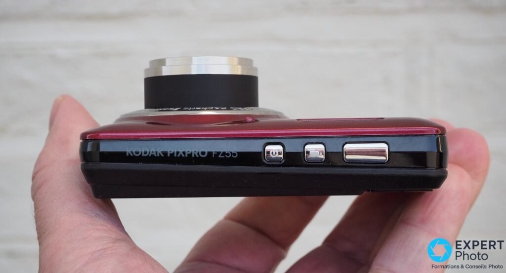 Tranche supérieure du Kodak Pixpro FZ55 rouge tenu en main, nom du modèle gravé, boutons et objectif déployé 