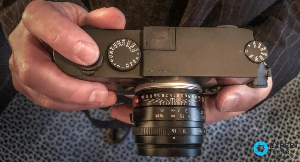 Leica M10 Monochrom tenu à deux mains, mollette ISO et molette vitesse visibles, optique 35 mm.