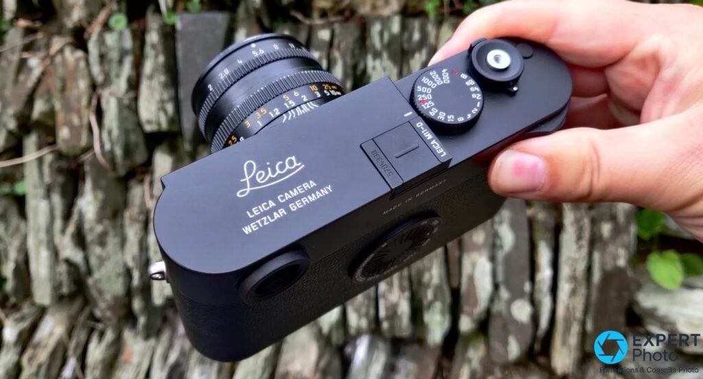 Boîtier Leica M11-D tenu en main vu en trois-quarts avant avec un objectif Leica M 50 mm monté, devant haie. 