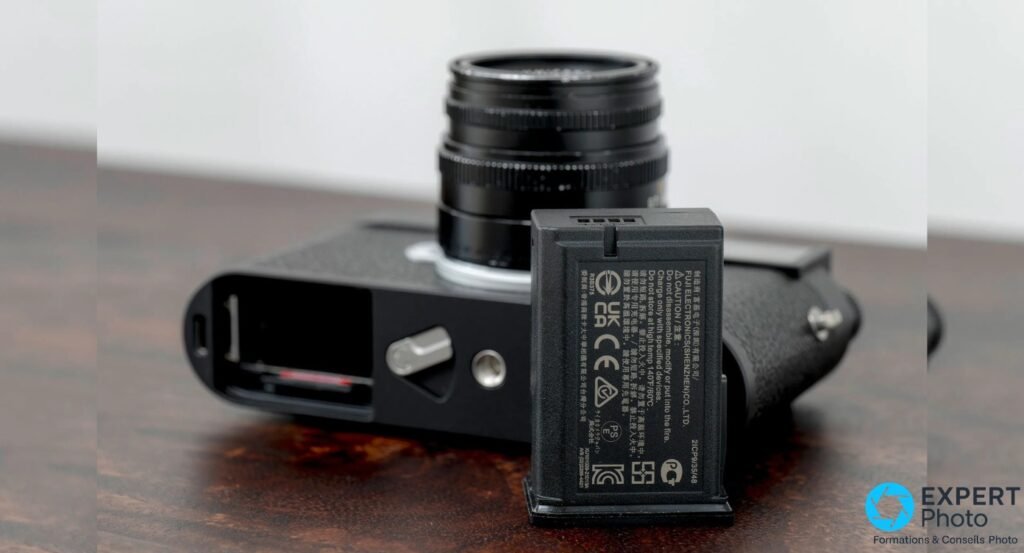 Leica M11-P noir avec trappe batterie ouverte et batterie extraite posée sur le dessus du boîtier, objectif partiellement visible
