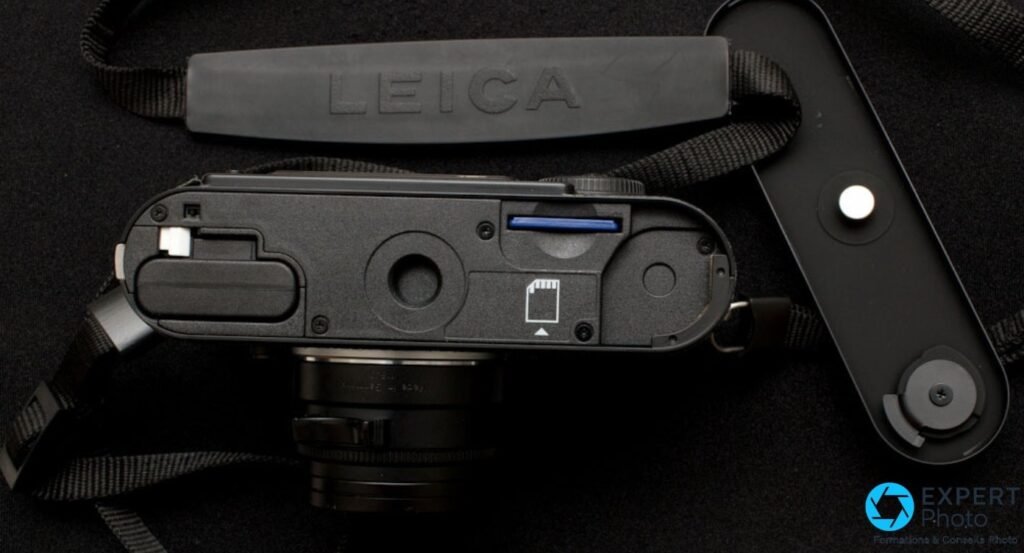 Semelle du Leica M9-P ouverte sur fond noir, logement de batterie et carte SD bleue inseree, dragonne Leica