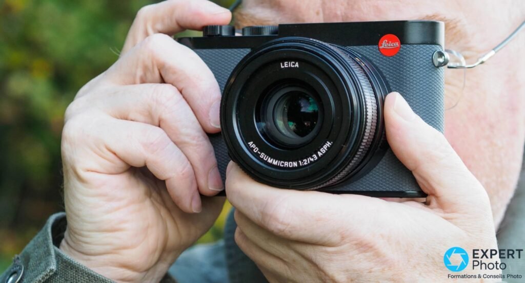 Leica Q3 43 gris tenu en position de visée, objectif APO-SUMMICRON 1:2/43 face caméra, fond végétation floue 