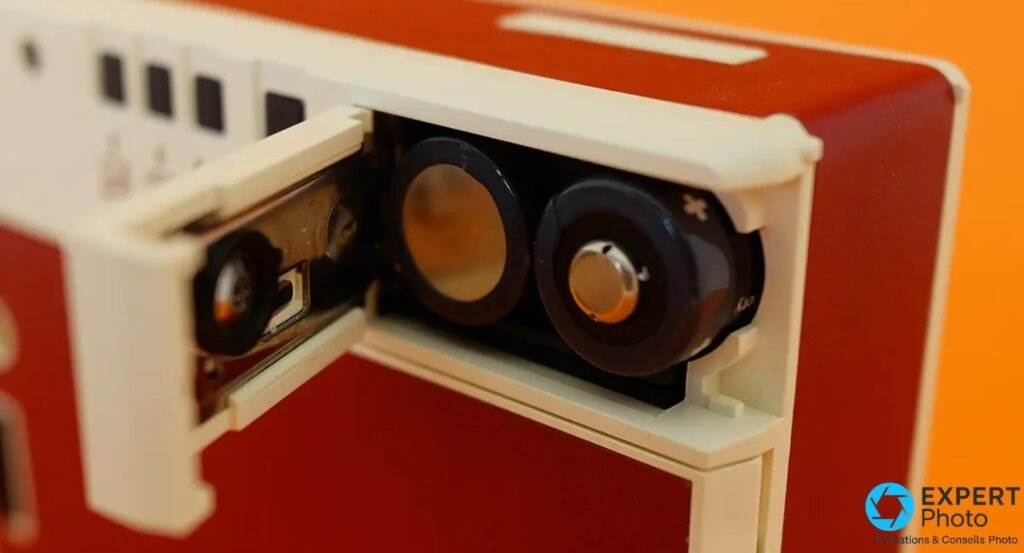 Lomo'Instant Automat, compartiment d'alimentation ouvert montrant deux piles rondes, fond orange