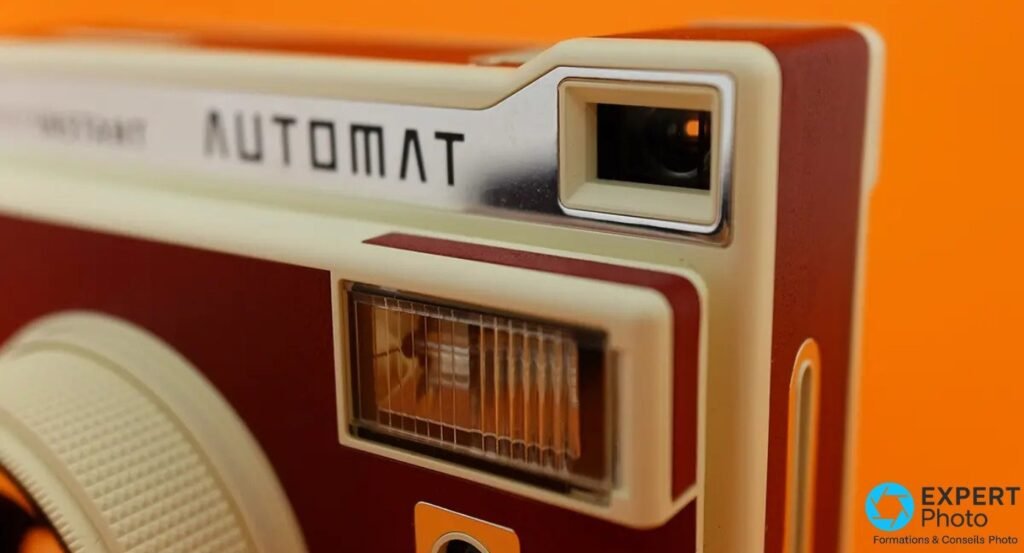 Lomo'Instant Automat, gros plan du viseur optique et du flash sous la gravure AUTOMAT, fond orange