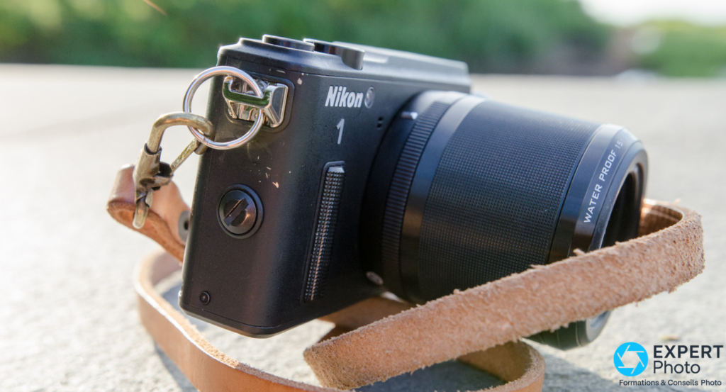 Nikon 1 AW1 noir vu de profil gauche, objectif 1 Nikkor AW monté, courroie cuir camel avec mousqueton, fond végétation