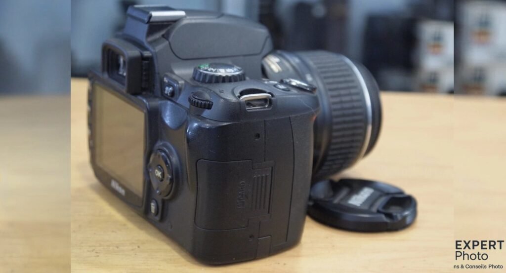 Nikon D40 en trois-quarts arrière droite, écran éteint, trappe carte SD visible, bouchon d'objectif posé à côté. 