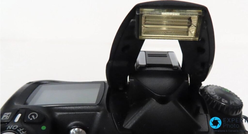 Gros plan du flash intégré du Nikon D50 relevé, lampe fresnel visible, bras articulé et griffe porte-flash. 